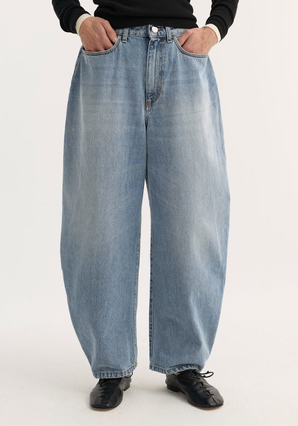 Sculptural denim trousers with balloon legs and high waist, worn with hands in pockets, showcasing a modern silhouette paired with a black rib-knit top.