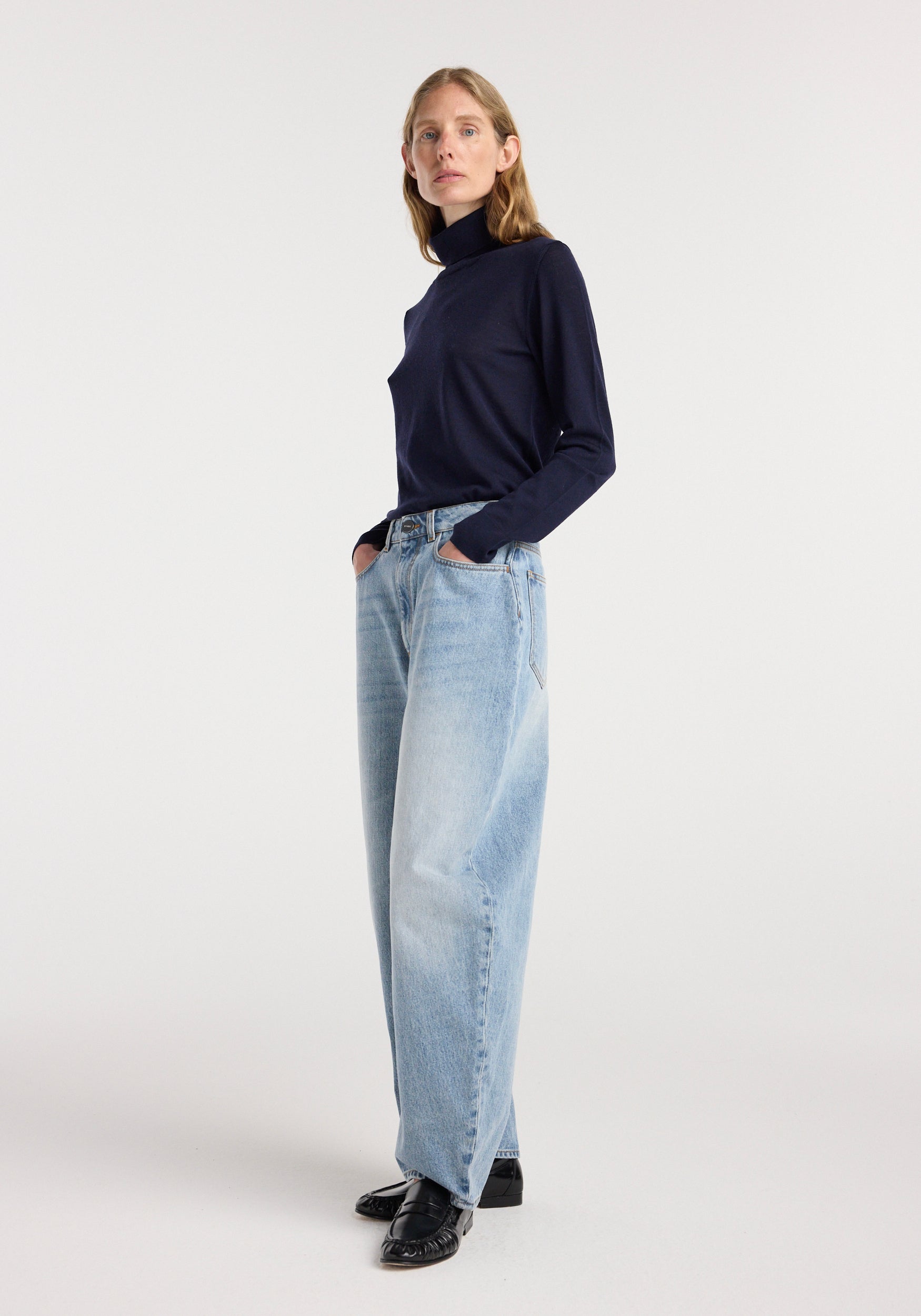 Woman wearing classic blue Fransje - Sculptural denim trousers with a high waist and wide leg, paired with a navy turtleneck and black shoes against a plain white background.