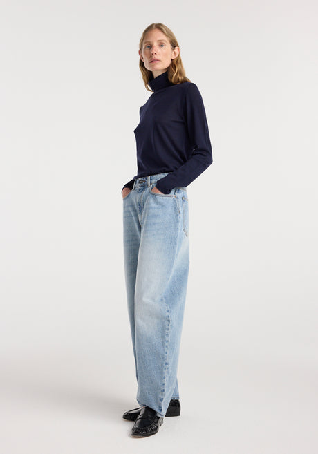 A woman wearing Fransje - Sculptural denim trousers | classic blue with a high waist and wide legs, paired with a fitted black turtleneck and black shoes, standing against a plain white background.