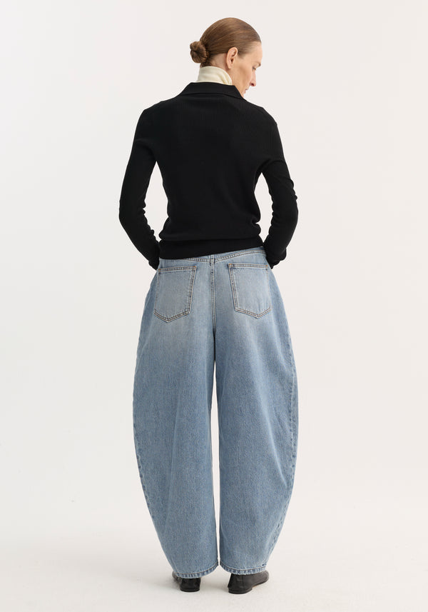 Model in sculptural denim trousers, classic blue; high-waisted, balloon leg design, paired with a slim black top, layered white collar, and black boots.