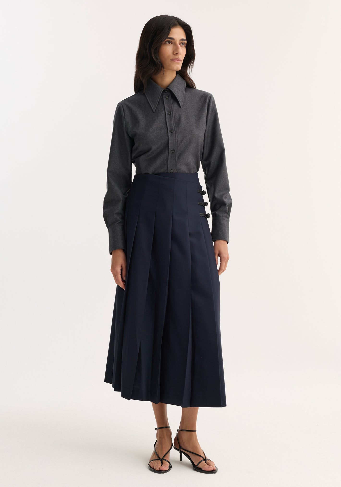 Woman wearing Pleated wool skirt with leather closures, featuring high-waisted fit, wide knife pleats, and decorative hip fastenings, paired with a charcoal-gray button-up shirt.