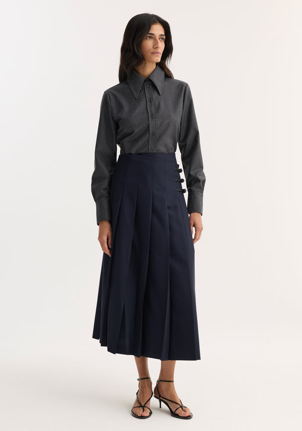 Woman wearing Pleated wool skirt with leather closures, featuring high-waisted fit, wide knife pleats, and decorative hip fastenings, paired with a charcoal-gray button-up shirt.