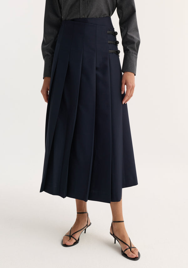 Woman wearing a navy Pleated wool skirt with leather closures, featuring wide pleats, fitted hip, and pankou closures, paired with a charcoal shirt and sandals.