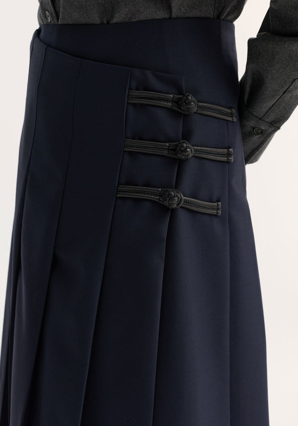 Pleated wool skirt with leather closures in navy, featuring high-waisted design, fitted hip, sharp pleats, and black frog-style toggle closures on the side.