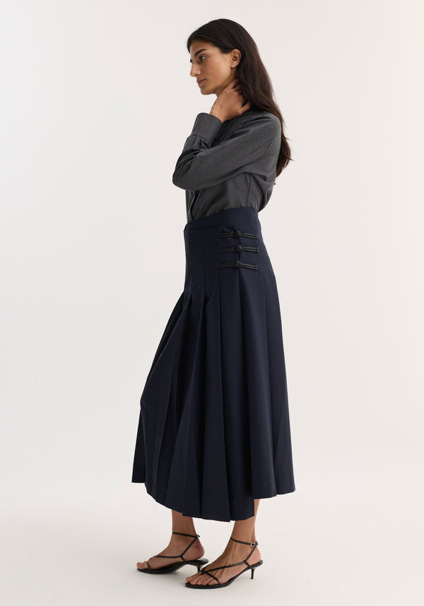 Woman in a Pleated wool skirt with leather closures, navy; fitted hip, box pleats, paired with a dark gray button-up shirt and black sandals.