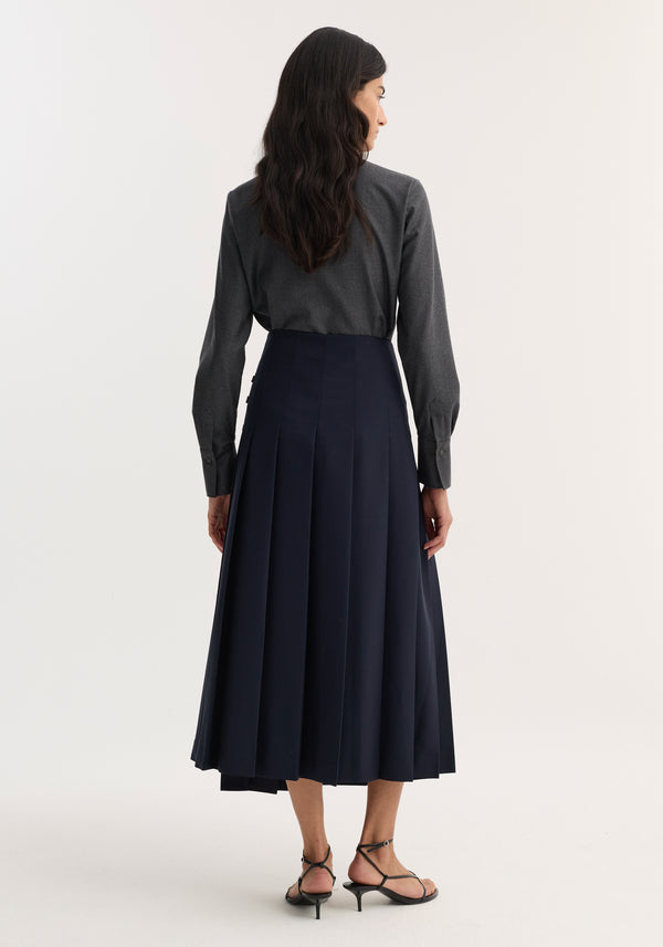 Rear view of a model wearing a Pleated wool skirt with leather closures, featuring a fitted hip and wide pleats, paired with a dark gray shirt and sandals.