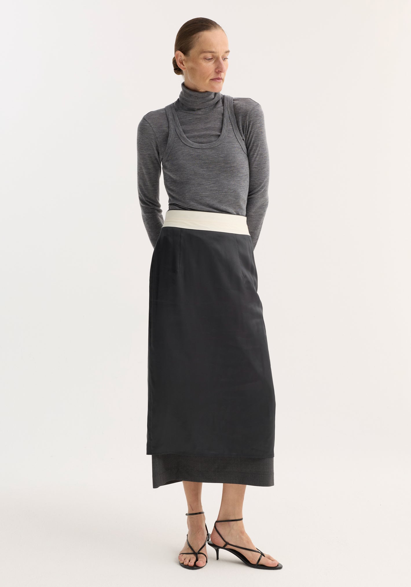 Model in Double-layer wool top | mid grey melange, showcasing a turtleneck under a tank, paired with a high-waisted skirt and strappy sandals.