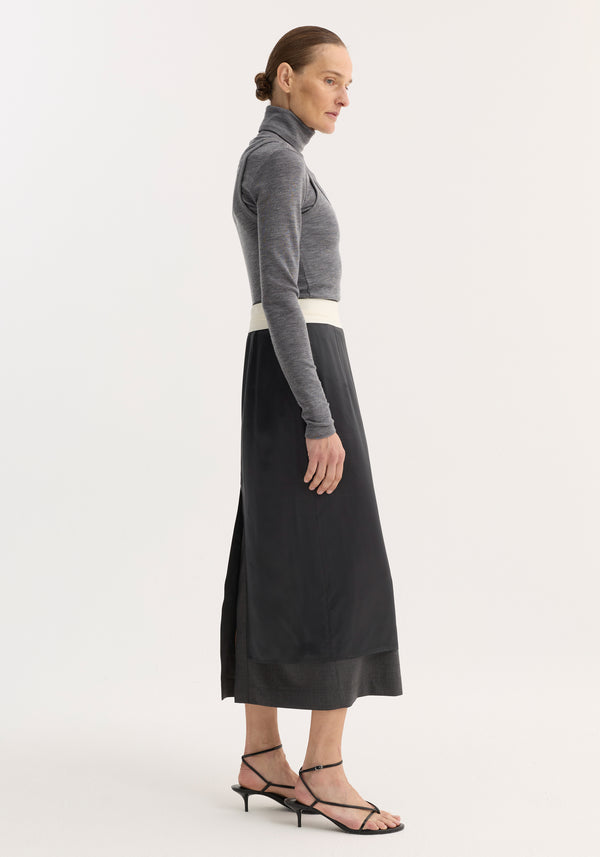 Woman models Inside-out skirt in dark grey melange, showcasing contrast waistband and side slit over darker underlayer, paired with a charcoal turtleneck and low-heel sandals.