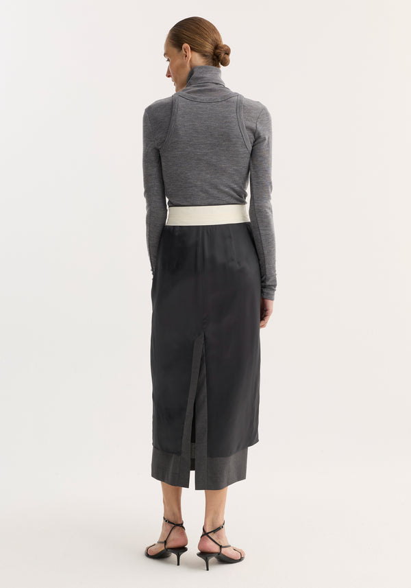 Rear-view of model in a Double-layer wool top, mid grey melange, showcasing a fitted turtleneck under a black apron-style skirt, with cropped trousers and sandals.