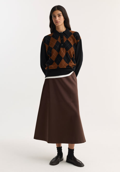 Woman wearing a voluminous A-line skirt in espresso satin, paired with an argyle sweater, black crew socks, and leather loafers, standing with hands behind her back.