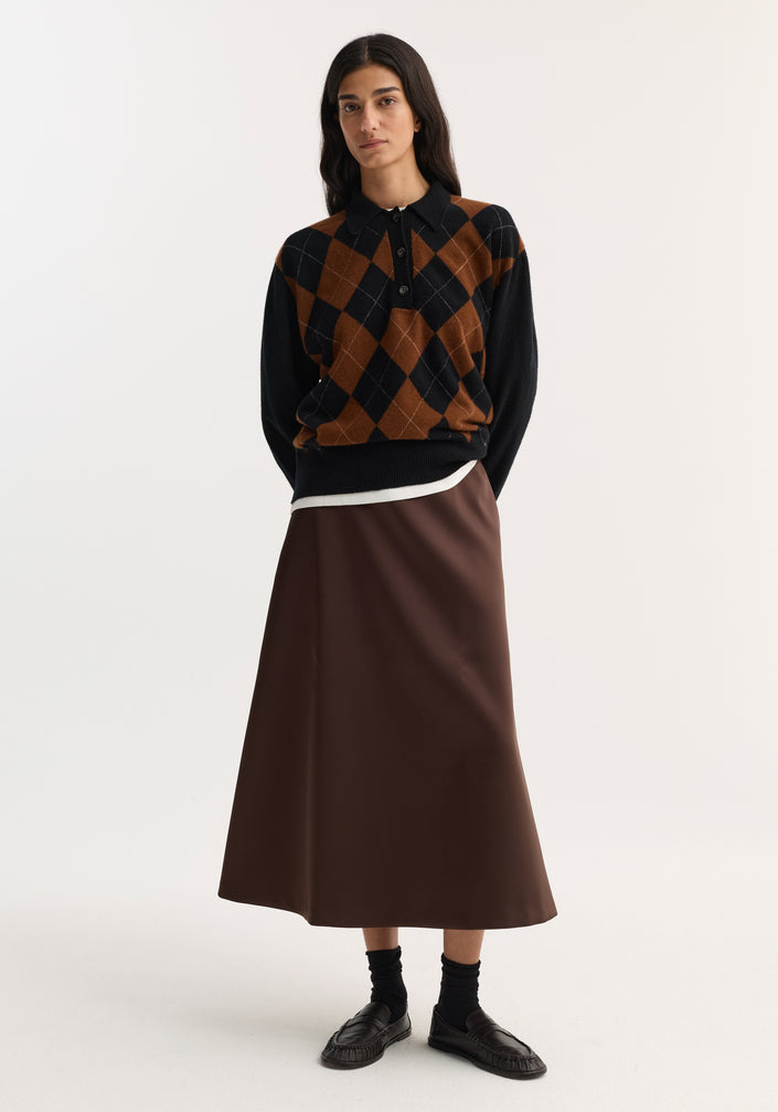 Woman wearing a voluminous A-line skirt in espresso satin, paired with an argyle sweater, black crew socks, and leather loafers, standing with hands behind her back.