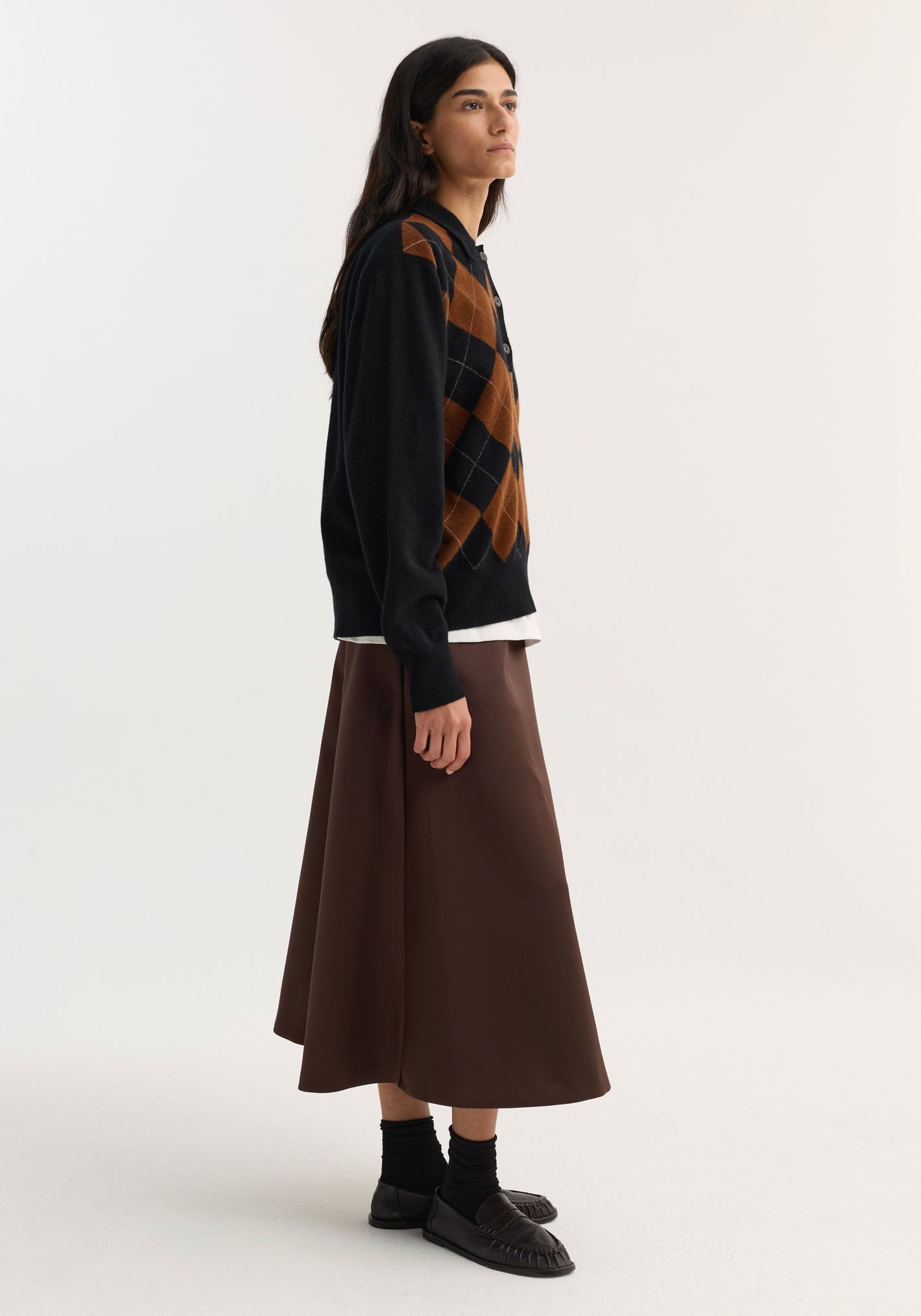 Model in profile wearing a voluminous A-line skirt in espresso satin, styled with a black argyle cardigan and black penny loafers.