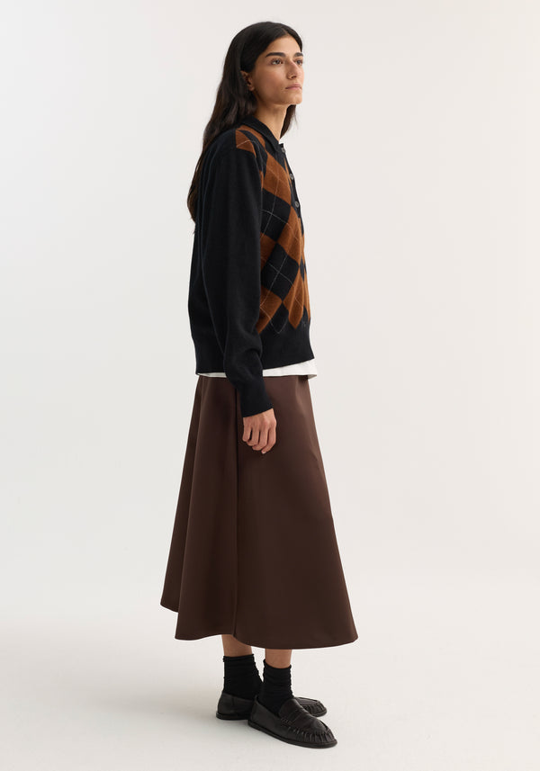 Model in profile wearing a voluminous A-line skirt in espresso satin, styled with a black argyle cardigan and black penny loafers.