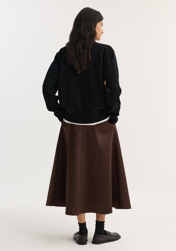Woman wearing a voluminous A-line skirt in espresso, featuring a full silhouette, over a white top and black sweater, with black shoes.
