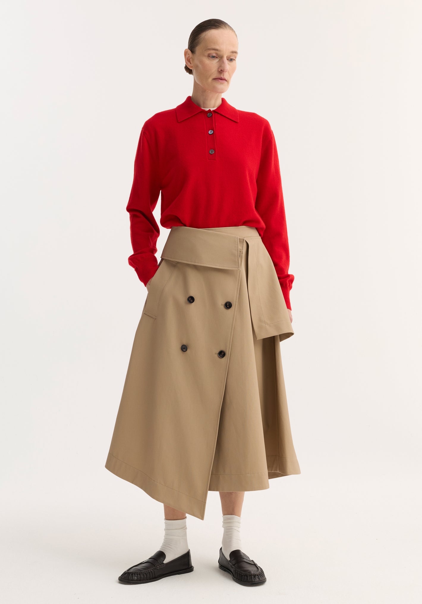 Model in a Deconstructed trench skirt | sand, featuring an asymmetrical wrap design, double-breasted buttons, and oversized belt flap, styled with a red polo sweater and black loafers.