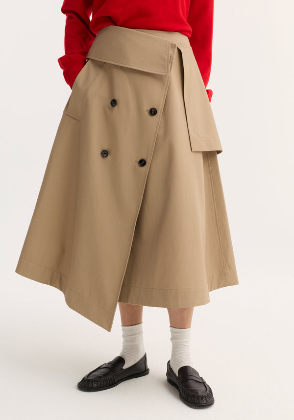 Deconstructed trench skirt in sand with asymmetric wrap, double-breasted buttons, oversized flap at waist, crafted from Japanese cotton. Paired with red top, socks, and loafers.