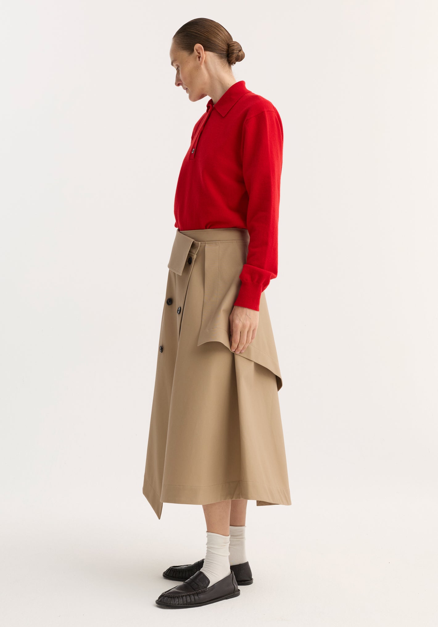 Side-profile of a model wearing a Deconstructed trench skirt | sand, featuring an asymmetrical flap, black buttons, and single side-seam pocket, paired with a red sweater.