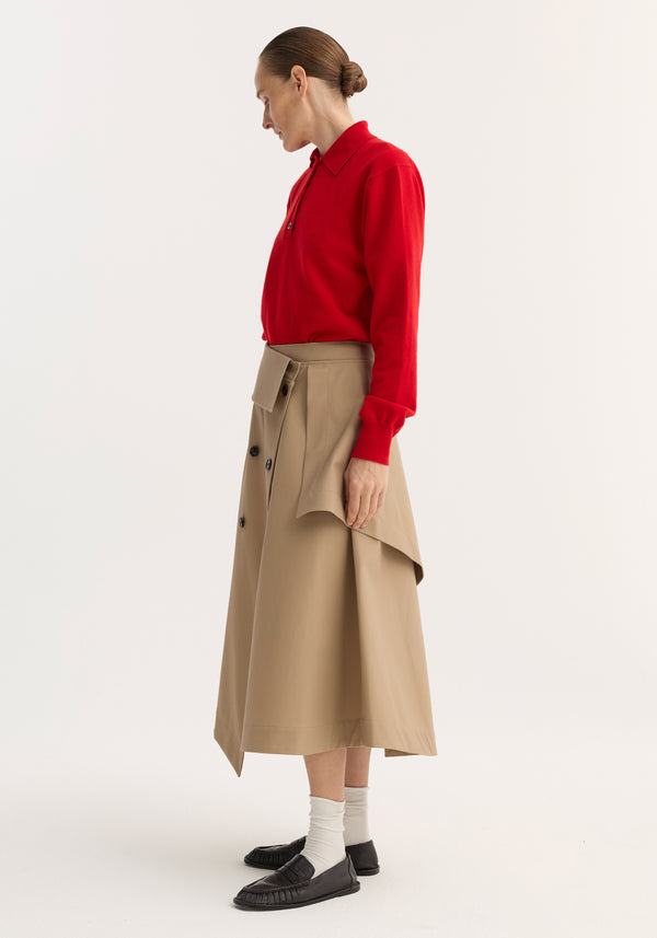 Side-profile of a model wearing a Deconstructed trench skirt | sand, featuring an asymmetrical flap, black buttons, and single side-seam pocket, paired with a red sweater.