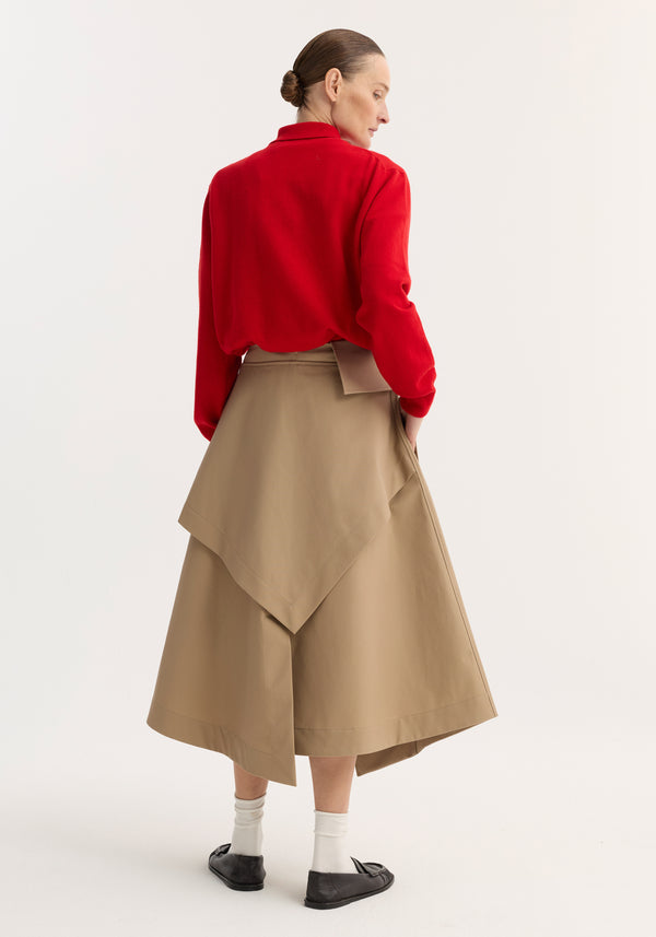 Person wearing a Deconstructed trench skirt | sand, featuring asymmetric hem, button details, and side pocket, paired with a red top and black slip-on shoes.