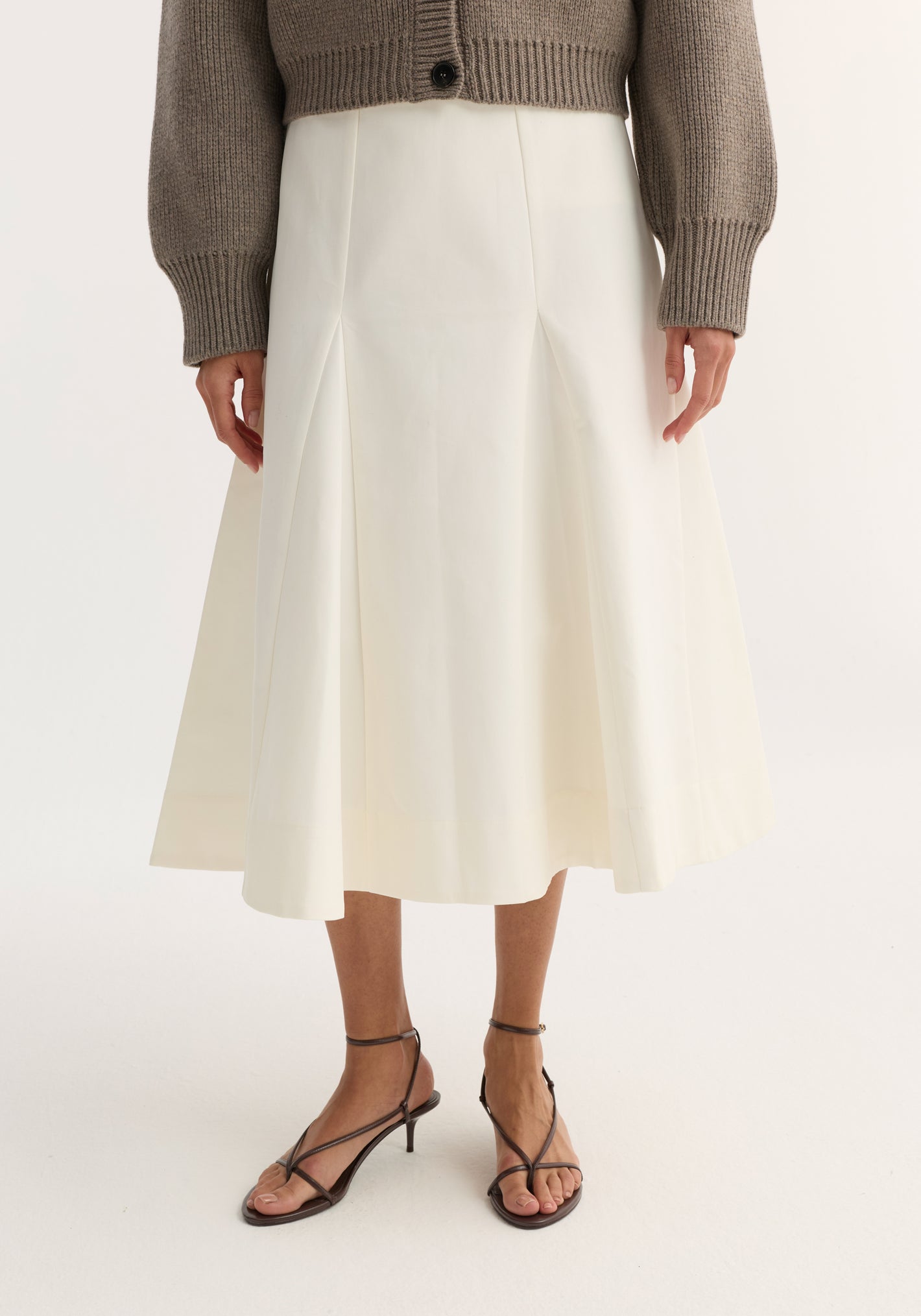 Sculptural cotton skirt in paper white, featuring a high-waisted A-line silhouette with structured pleats, paired with black sandals and a taupe cardigan.