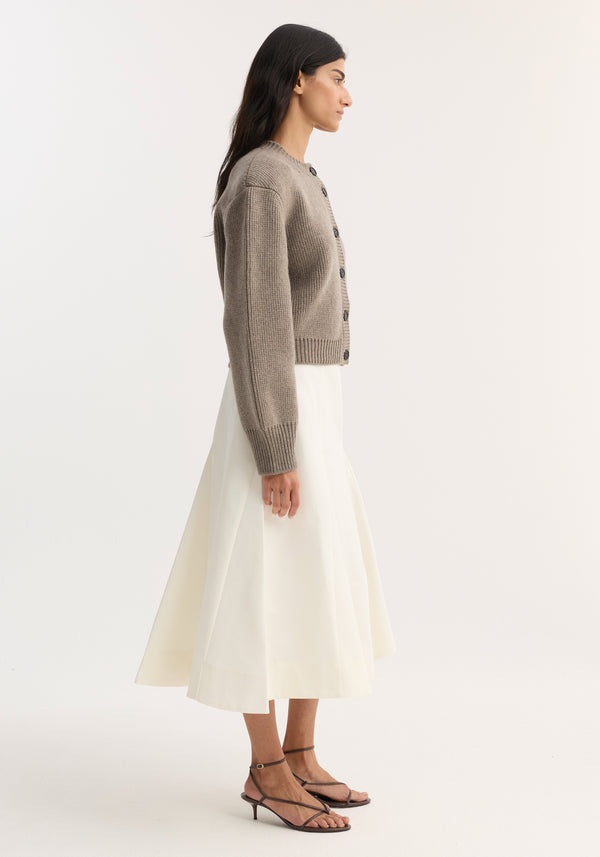 Model in a Sculptural cotton skirt | paper white, featuring a high-waisted, A-line silhouette with structured pleats, paired with a taupe cardigan and black sandals.