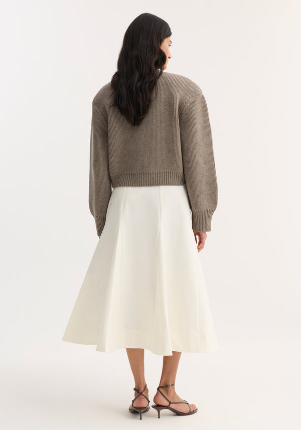 A woman models the Sculptural cotton skirt in paper white, featuring a fitted waist and A-line silhouette with structured pleats, paired with a taupe knit sweater.