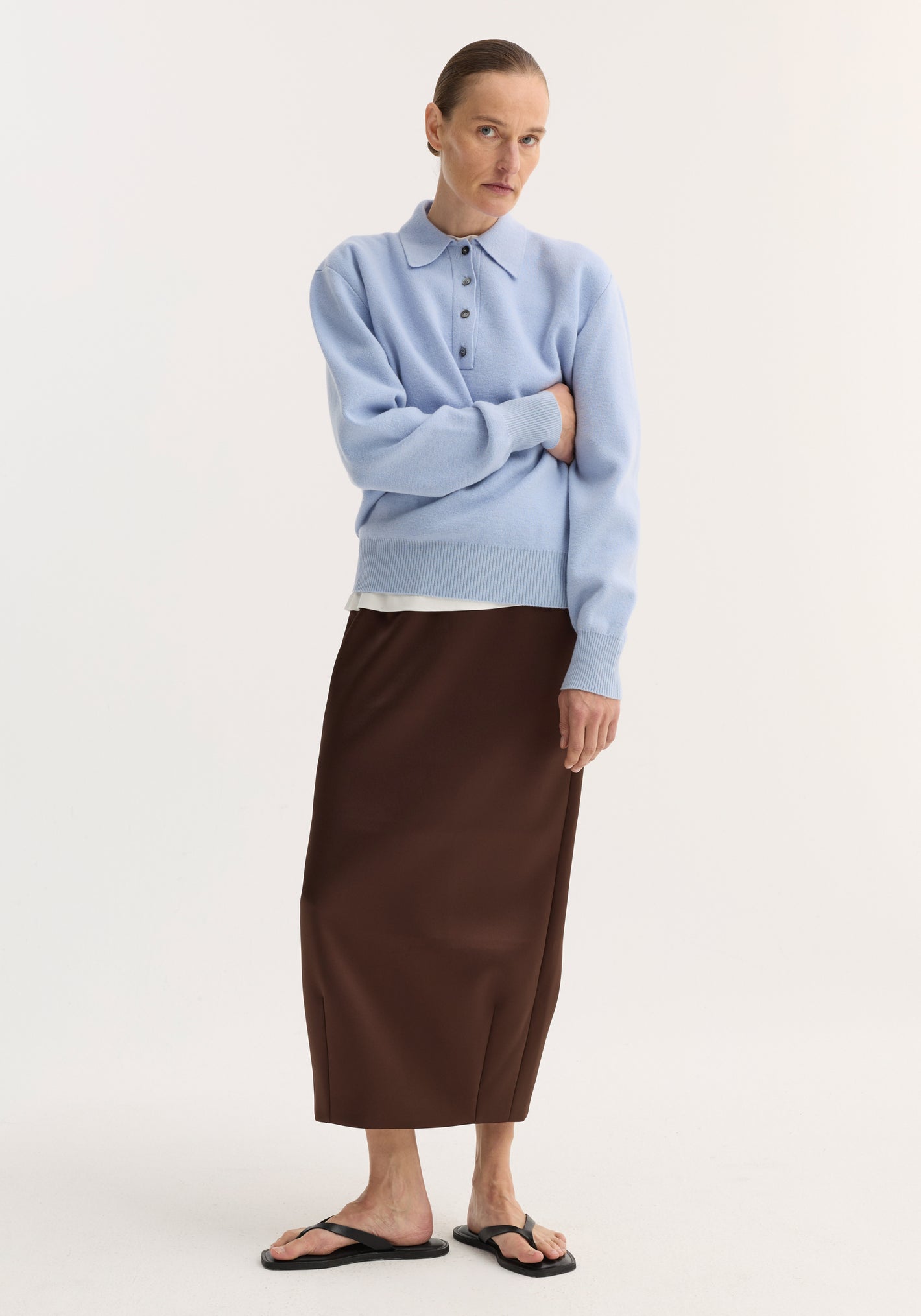 Woman wearing a Balloon column skirt | espresso, high-waist with dart detailing, paired with a light-blue knit polo sweater and black flip-flops, against a plain backdrop.