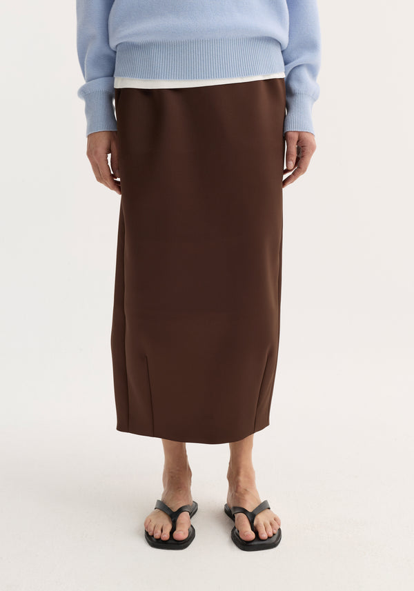 Balloon column skirt in espresso with a sculptural cut, high waist, dart detailing, and a back slit, worn with a knit sweater and sandals.
