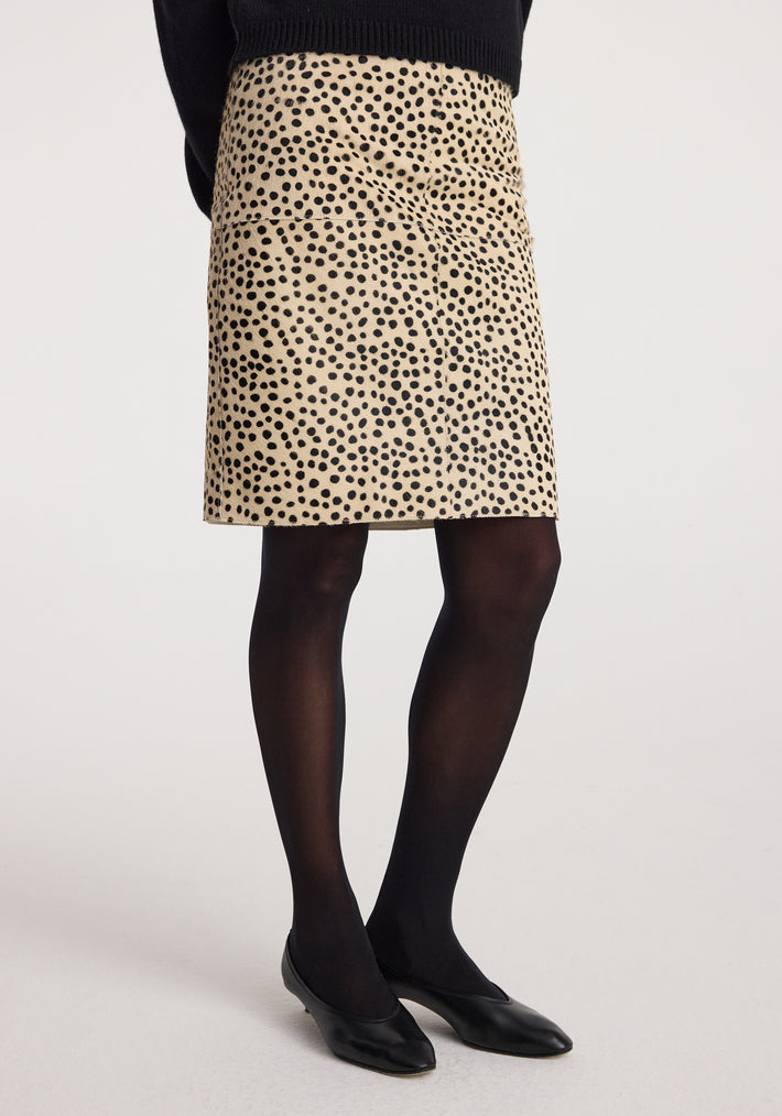 Woman wearing a beige Pony hair pencil skirt | cheetah with black spot pattern, paired with sheer black tights and black flat shoes against a plain background.