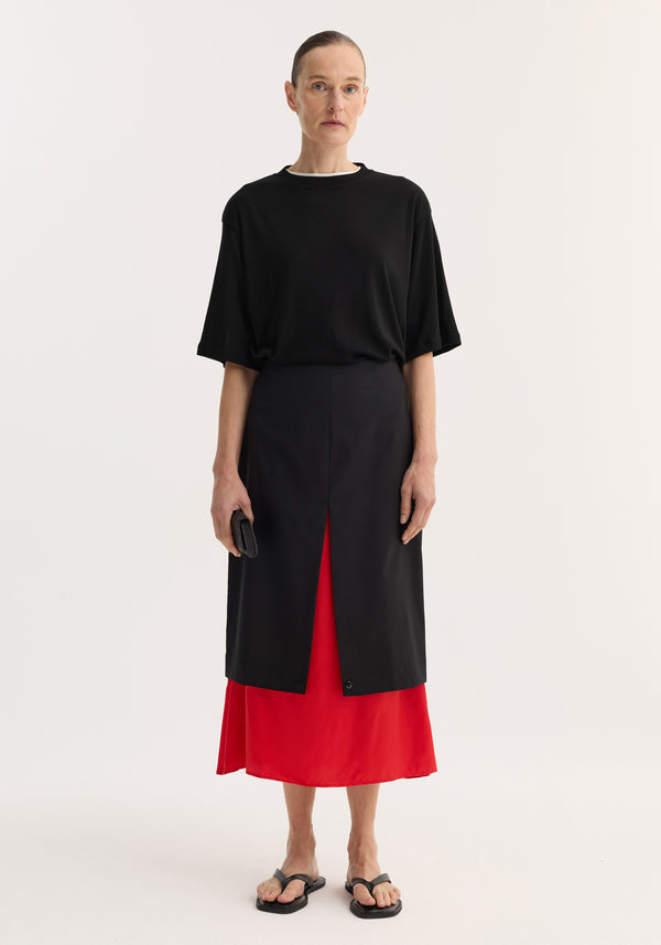 Model in a Contrast upside-down skirt | black featuring a black pencil skirt with red underlay, front slit, black top, and flip-flop sandals, holding a clutch.