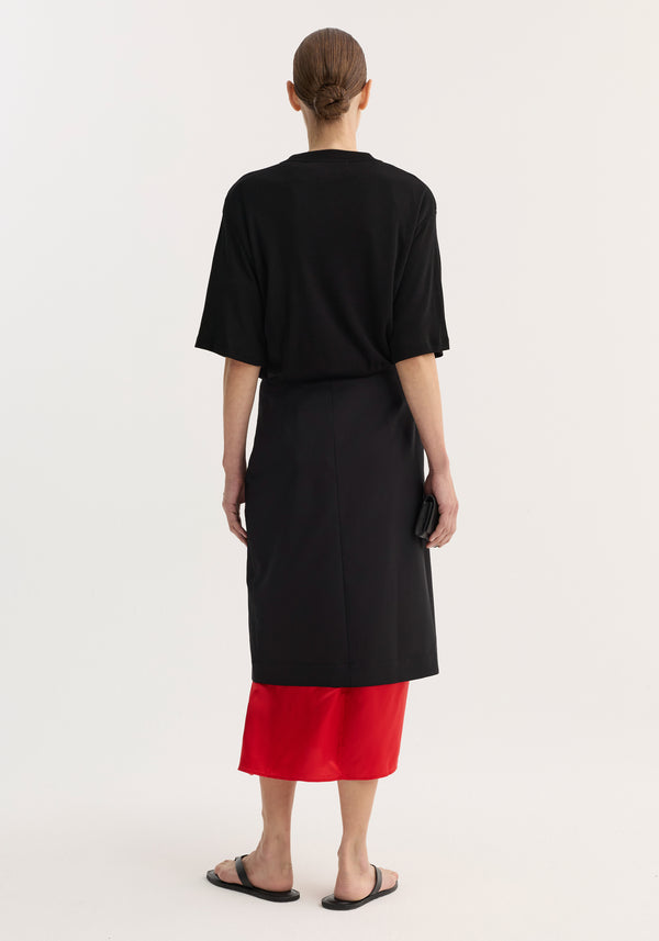 Woman wearing Contrast upside-down skirt | black with tapered silhouette, front slit, and bold red underlayer, paired with a loose black top and black sandals.