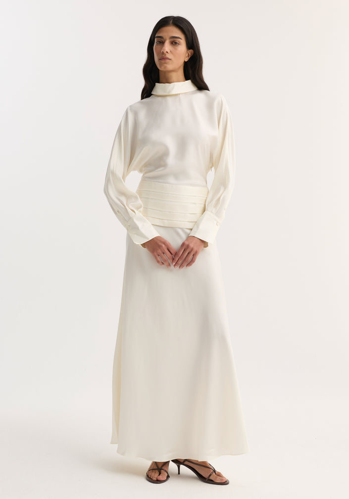 Model in Tuxedo silk dress | cream, featuring a high-neck blouse, pleated waistband, A-line skirt, and open back with cummerbund and side zip detailing.