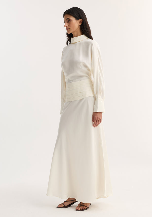 Model wearing Tuxedo silk dress | cream, featuring flowing sleeves, high collar, cummerbund detailing, open back, and side zip, paired with black strappy sandals.