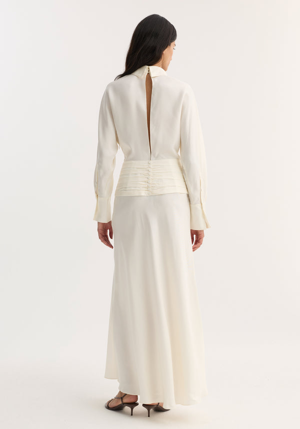 Tuxedo silk dress | cream: Woman in a long-sleeved silk dress with cummerbund waist, open back, high collar, and pleated skirt, wearing strappy sandals.