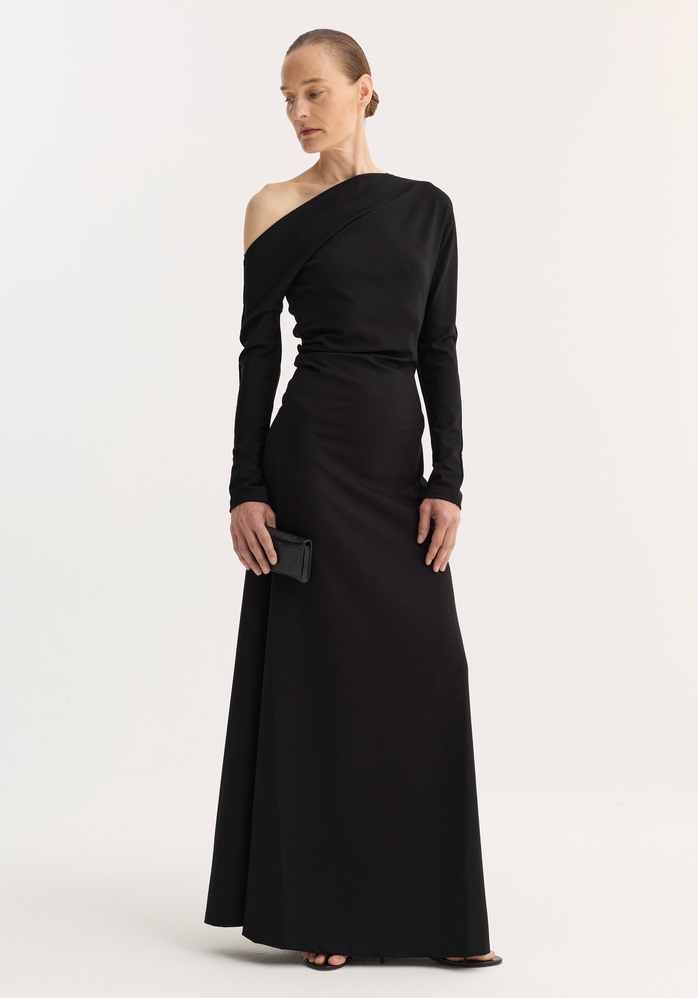 Woman in a Twisted jersey dress | black, featuring an off-the-shoulder neckline, long sleeves, and a gathered waist, holding a black clutch.