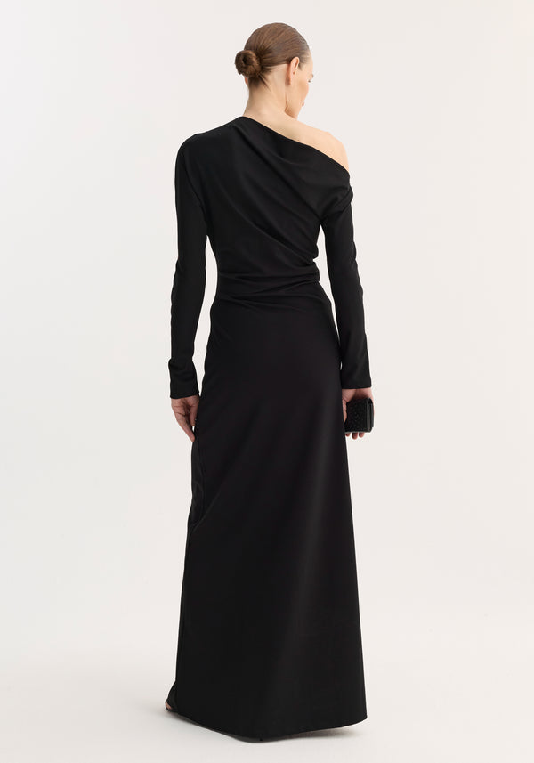 A woman in a floor-length Twisted jersey dress | black, featuring an asymmetric neckline and long sleeves, holds a black clutch with her hair in a bun.