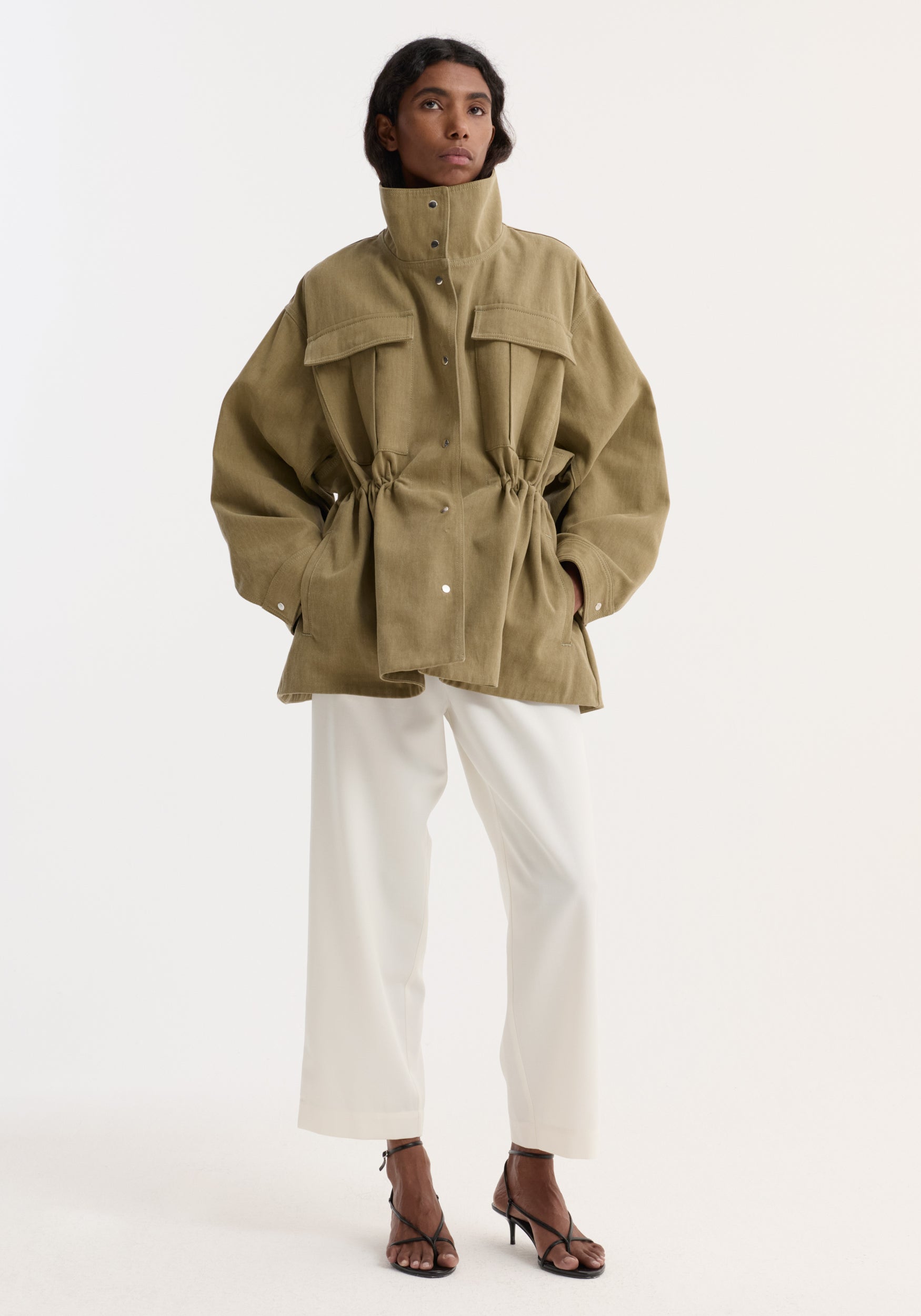Woman wearing Sculptural safari jacket | sage, featuring a high collar, oversized pockets, cinched waist. Paired with wide-leg trousers and strappy heels against a neutral backdrop.