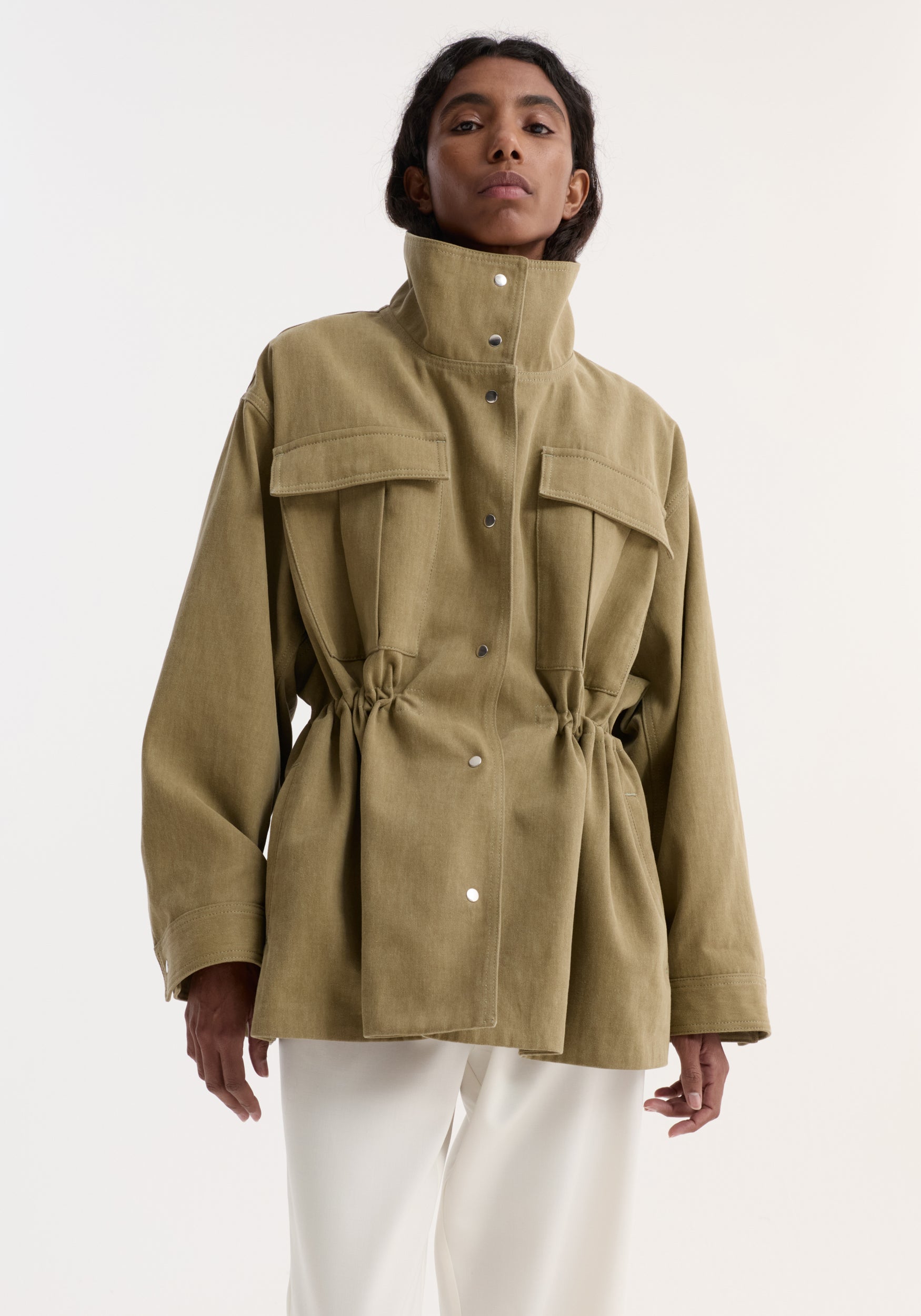 Model wearing Sculptural safari jacket | sage, featuring a high snap-close collar, large flap chest pockets, and adjustable drawstring waist, paired with loose white trousers.