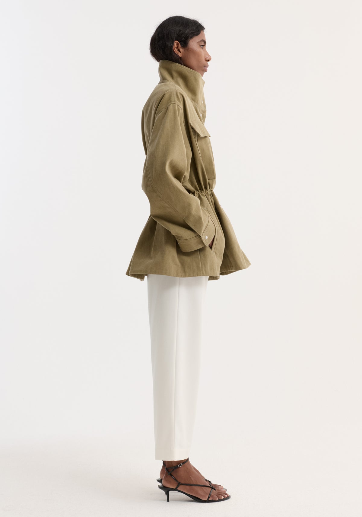 Model in profile wearing a Sculptural safari jacket | sage with a stand collar, bellows pockets, and elasticated waist, paired with slim white trousers and black sandals.