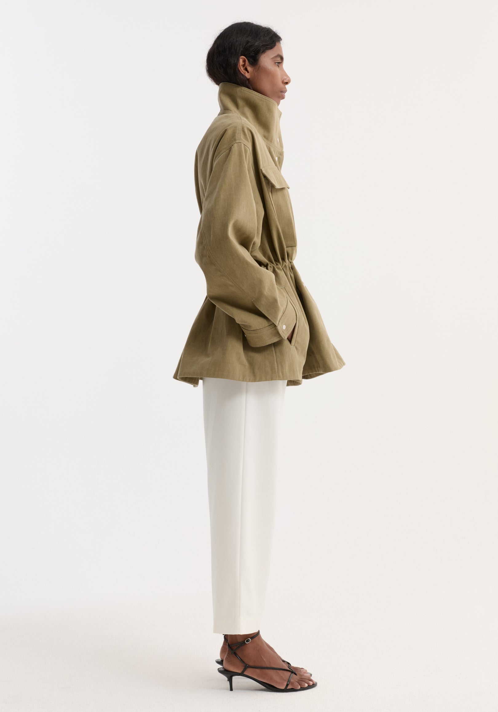 Model in profile wearing a Sculptural safari jacket | sage with a stand collar, bellows pockets, and elasticated waist, paired with slim white trousers and black sandals.