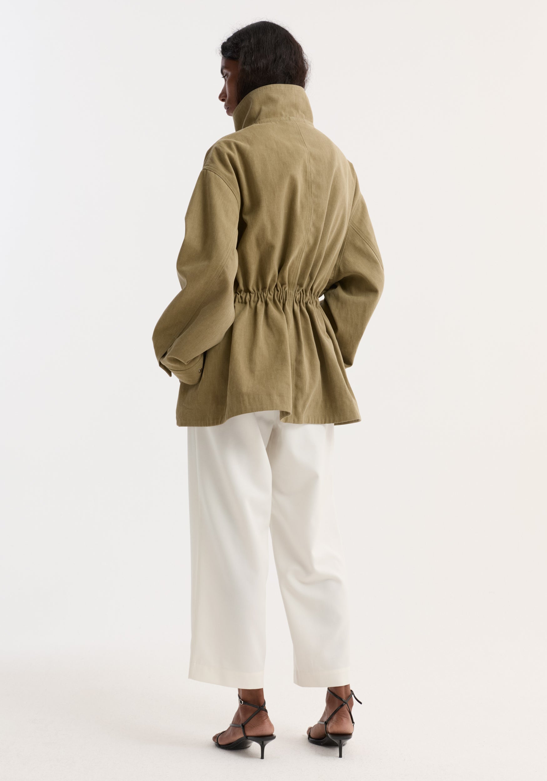 Woman in a Sculptural Safari Jacket | Sage, showing a high collar and elastic waist flare, paired with wide-leg trousers and strappy sandals.