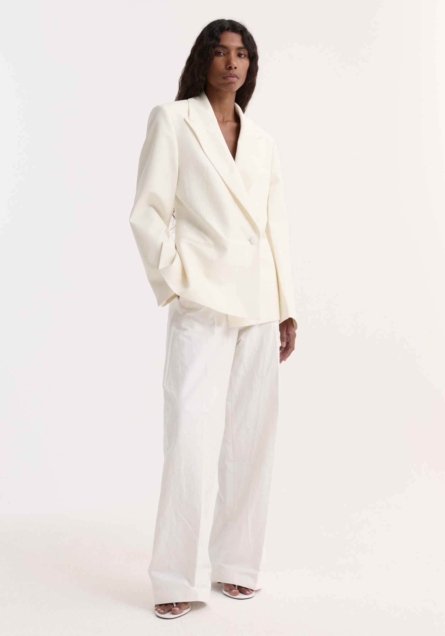 Model wearing Hourglass shaped blazer in off white, featuring sharp notch lapel, cinched waist, paired with wide-leg trousers and open-toe heels.