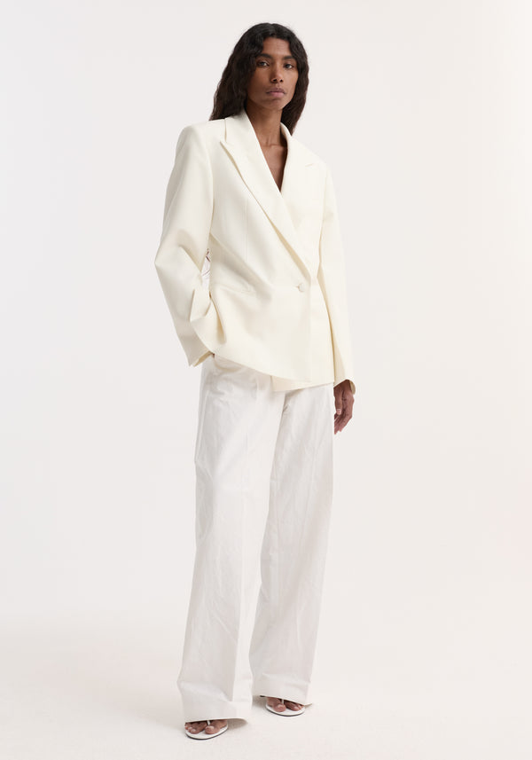 Model wearing Hourglass shaped blazer in off white, featuring sharp notch lapel, cinched waist, paired with wide-leg trousers and open-toe heels.