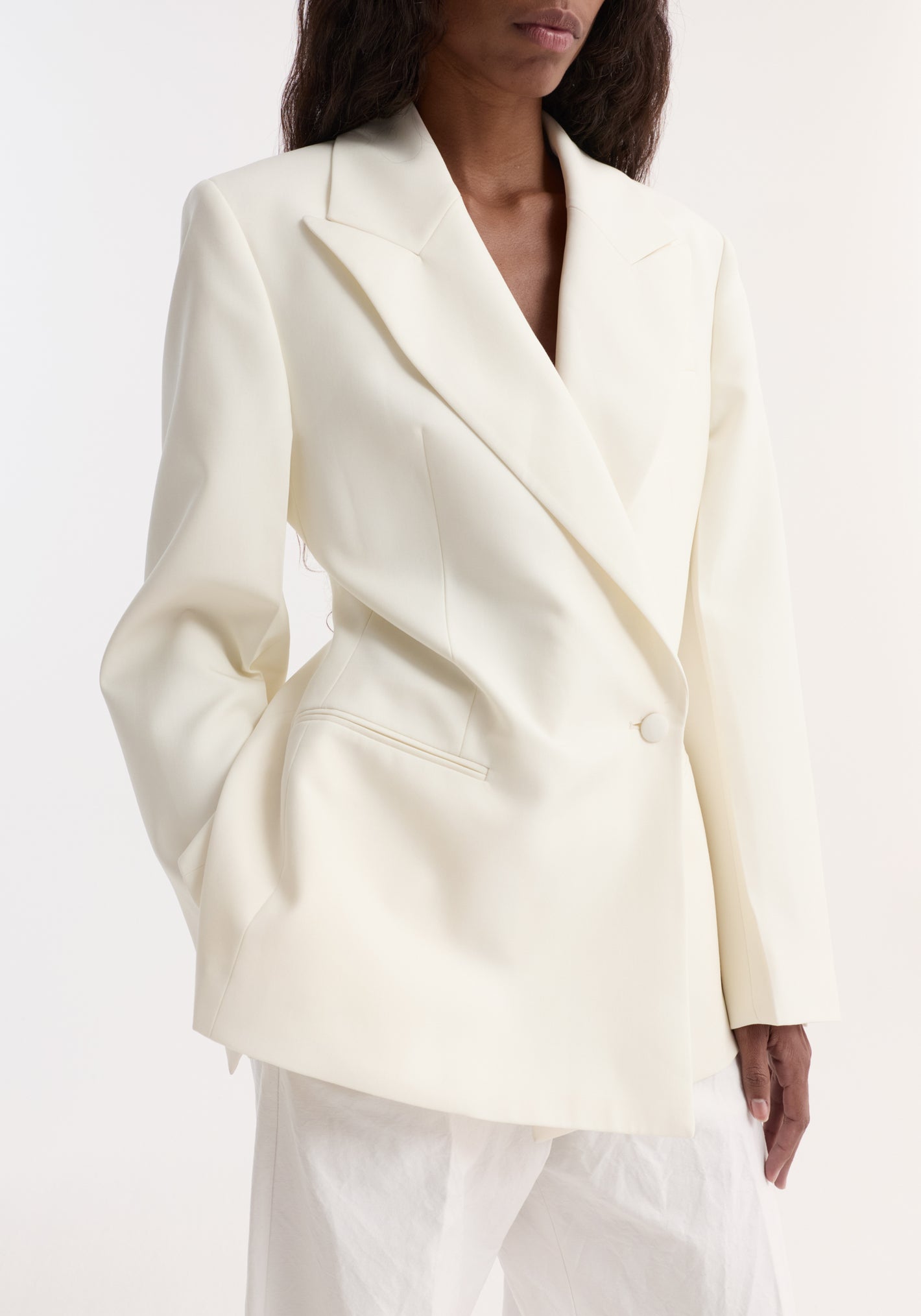 Model wears an Hourglass shaped blazer in off-white with a sharp notch lapel, cinched waist, single button closure, and welt pockets, paired with white trousers.