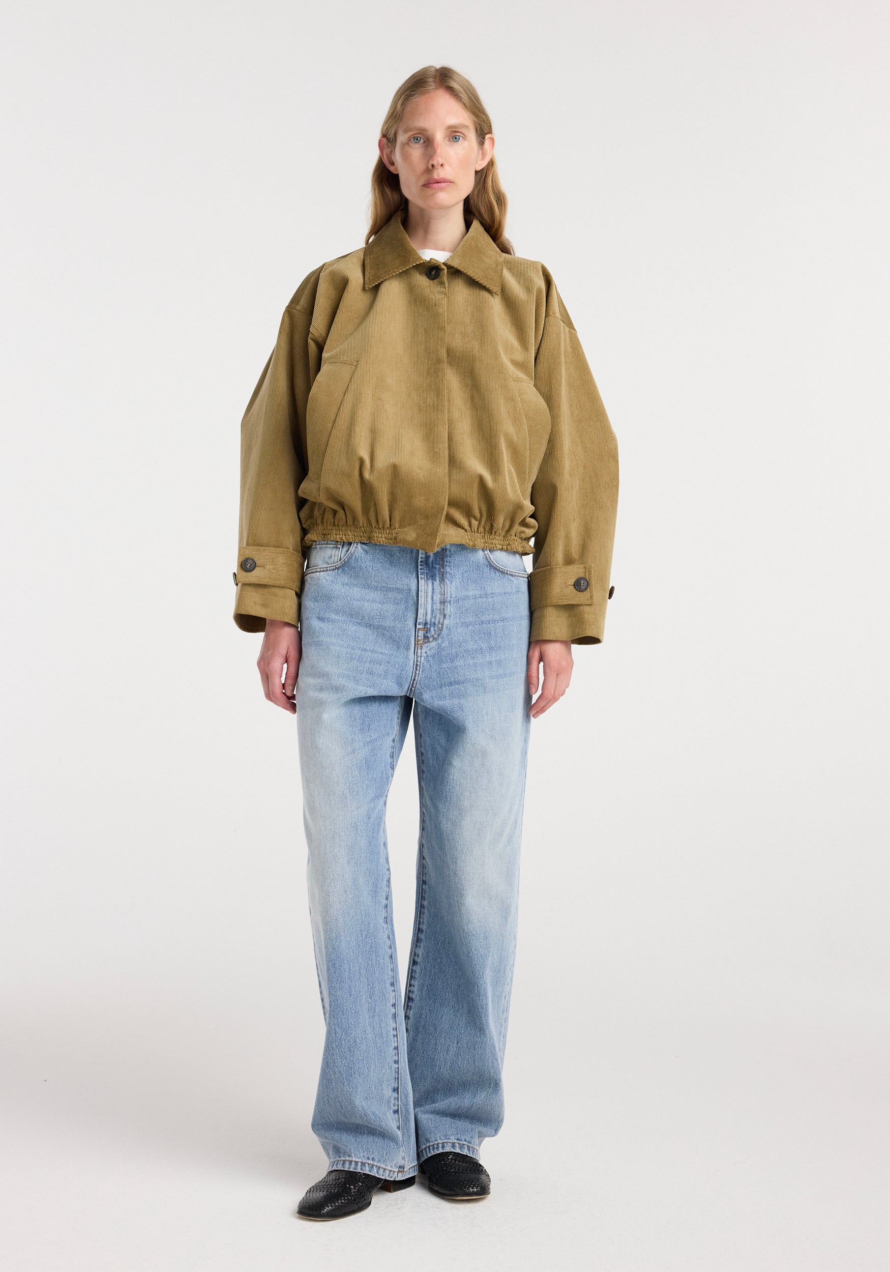 Front view of a woman wearing a beige Corduroy blouson jacket | beige with long sleeves, a collar, and buttoned cuffs paired with light blue wide-leg jeans and black shoes against a plain white background.