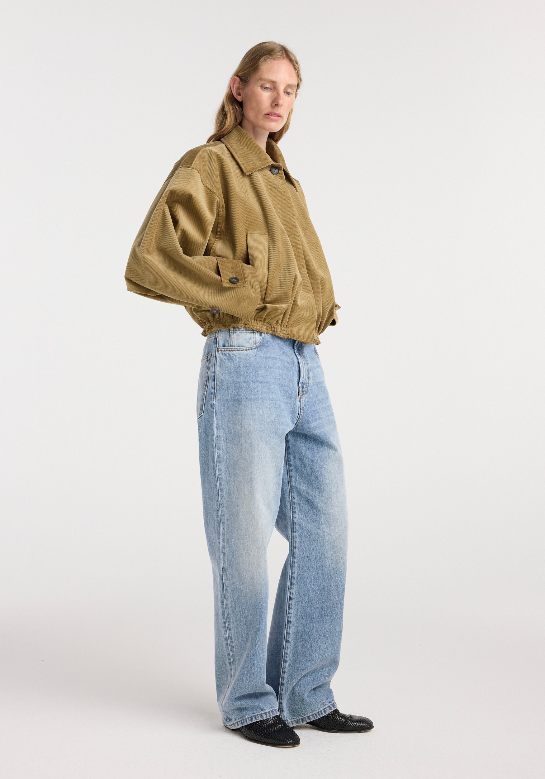 Woman wearing a beige Corduroy blouson jacket with wide sleeves and buttoned cuffs, paired with light blue denim jeans and black shoes, standing against a plain background.