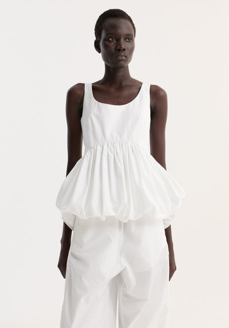 Model in Voluminous Peplum Top | Optic White, featuring a sleeveless scoop neck, low back, and bubble-hem waist, paired with loose, elastic-cuffed trousers.
