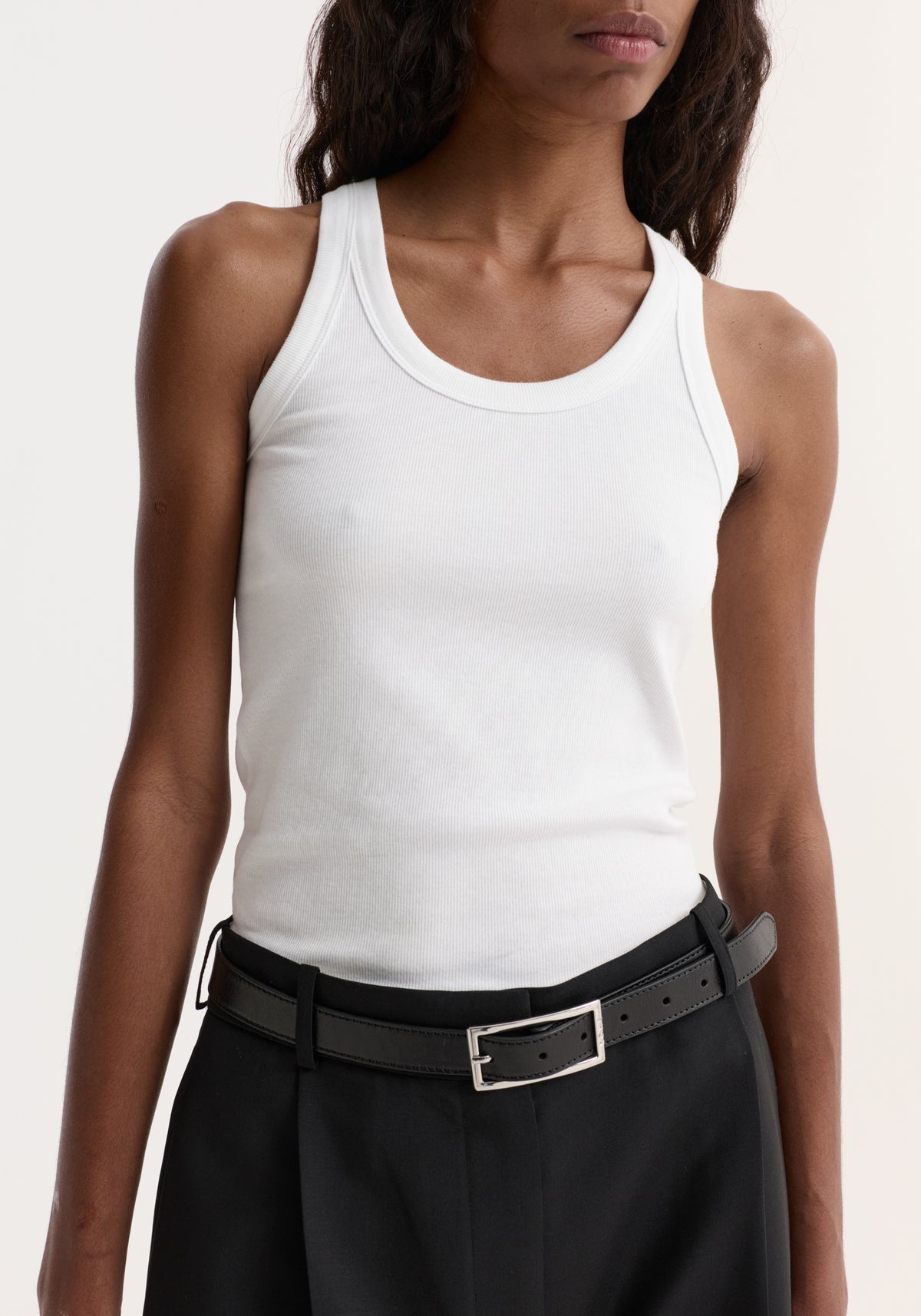 Person wearing a Scoop-neck rib top in optic white, featuring a scoop neck and racerback, paired with high-waisted black trousers and a slim leather belt.