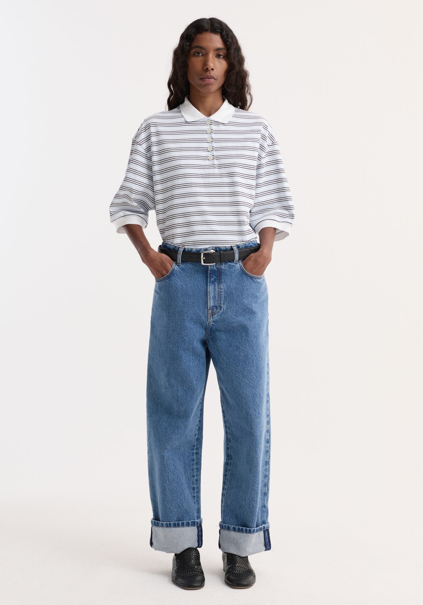 Person wearing a striped oversized polo with a white and blue multi-stripe design, high-waisted blue jeans, black belt, and black woven slip-on shoes.