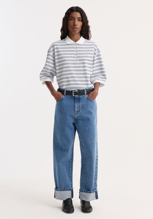 Person wearing a striped oversized polo with a white and blue multi-stripe design, high-waisted blue jeans, black belt, and black woven slip-on shoes.